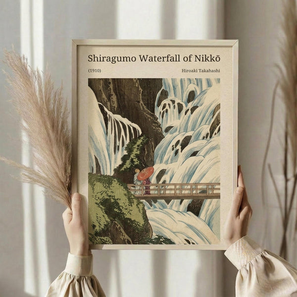 May include: Hands holding a framed vintage Japanese woodblock print depicting a dramatic waterfall cascading over rocky cliffs with a small figure holding a red umbrella on a wooden bridge. The artwork features muted blue and white water against dark rocks with green moss, displayed in a light wooden frame. Dried pampas grass stems are held alongside the frame against flowing white curtains in soft natural lighting.