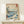 May include: Framed Japanese woodblock print reproduction featuring dramatic blue ocean waves, Mount Fuji in background, pine tree on rocky coastline, small sailing boat, and flying birds. Print displays in natural wood frame on light wooden surface with small wrapped gift box. Wall mount setup shows vintage ukiyo-e art style with traditional red text cartouches and muted color palette of blues, greens, and cream tones.