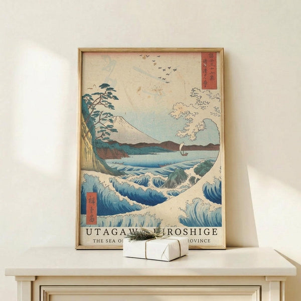 May include: Framed Japanese woodblock print reproduction featuring dramatic blue ocean waves, Mount Fuji in background, pine tree on rocky coastline, small sailing boat, and flying birds. Print displays in natural wood frame on light wooden surface with small wrapped gift box. Wall mount setup shows vintage ukiyo-e art style with traditional red text cartouches and muted color palette of blues, greens, and cream tones.