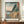 May include: Framed vintage Japanese woodblock print showing dramatic coastal cliffs with caves and crashing blue waves. Features Mount Fuji in background, traditional pagoda on clifftop, and pink-orange sunset sky. Print has Japanese text cartouches and sits on wooden credenza beside white ceramic vase and books in cream-walled room with white trim.