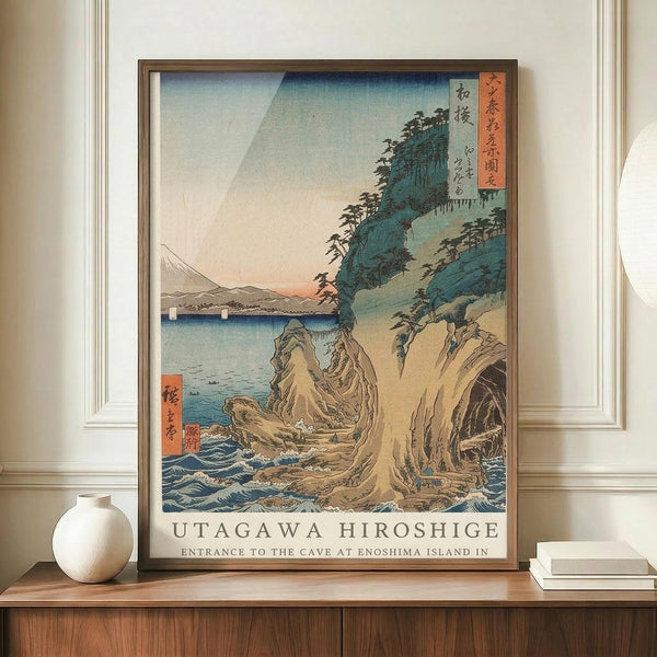 May include: Framed vintage Japanese woodblock print showing dramatic coastal cliffs with caves and crashing blue waves. Features Mount Fuji in background, traditional pagoda on clifftop, and pink-orange sunset sky. Print has Japanese text cartouches and sits on wooden credenza beside white ceramic vase and books in cream-walled room with white trim.