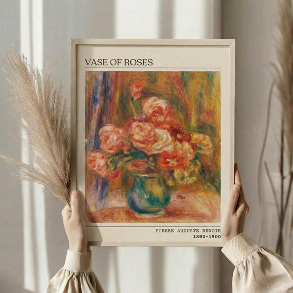 May include: Person in cream ruffled sleeve top holding framed art print against white curtained window, with dried pampas grass visible. Frame shows Impressionist-style painting of pink and coral roses in blue-green vase with warm golden background. Print has museum-style cream matting with "VASE OF ROSES" at top and "PIERRE AUGUSTE RENOIR 1890-1900" at bottom in black text.