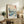 May include: Framed Japanese woodblock print featuring blue ocean waves, Mount Fuji, flying birds, and traditional red text cartouches, displayed on rustic wooden sideboard alongside fern plant, ceramic vase, and books in neutral-toned living room setting with beige chair and white walls.