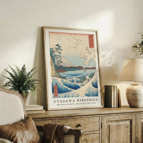 May include: Framed Japanese woodblock print featuring blue ocean waves, Mount Fuji, flying birds, and traditional red text cartouches, displayed on rustic wooden sideboard alongside fern plant, ceramic vase, and books in neutral-toned living room setting with beige chair and white walls.
