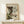 May include: A framed vintage Japanese woodblock print depicting a dramatic winter waterfall scene with cascading white water, dark rocky cliffs, green vegetation, and small figures on a wooden bridge. The artwork features traditional ukiyo-e style with blue and white water flows against brown rocks. Displayed in a light wood frame on a white wooden console table beside a small wrapped gift box, against a cream wall.