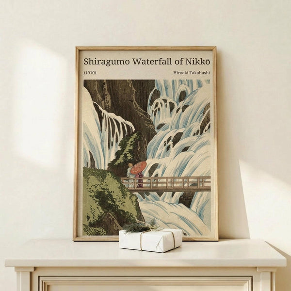 May include: A framed vintage Japanese woodblock print depicting a dramatic winter waterfall scene with cascading white water, dark rocky cliffs, green vegetation, and small figures on a wooden bridge. The artwork features traditional ukiyo-e style with blue and white water flows against brown rocks. Displayed in a light wood frame on a white wooden console table beside a small wrapped gift box, against a cream wall.