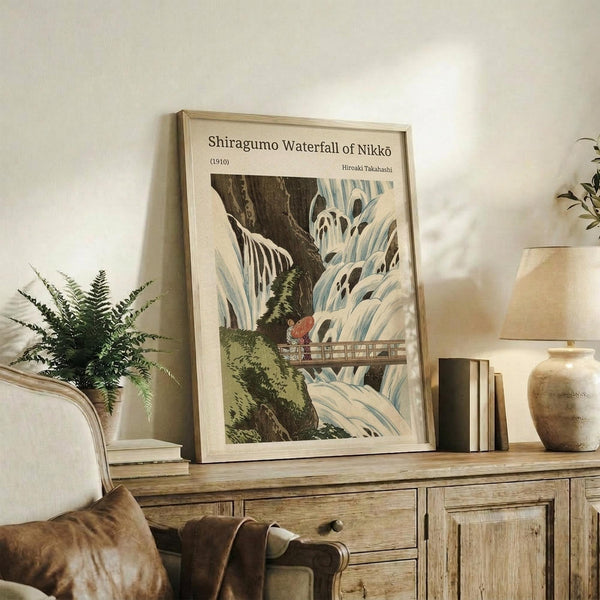 May include: Framed vintage Japanese woodblock print artwork showing a dramatic waterfall scene with flowing white water cascading over dark rocks, small figures on a wooden bridge, and green foliage. The beige-framed print sits on a rustic wooden sideboard alongside a fern plant, ceramic vase, and books in a neutral-toned living room with cream upholstered furniture.