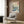 May include: Framed Japanese woodblock print featuring blue ocean waves, Mount Fuji, and birds in flight, displayed above a beige upholstered armchair with geometric pillow. Modern living room setting with brass floor lamp, dark round side table with open book, and wood-paneled wall. Print shows traditional ukiyo-e art style with vibrant blues, coral accents, and cream tones in white frame.