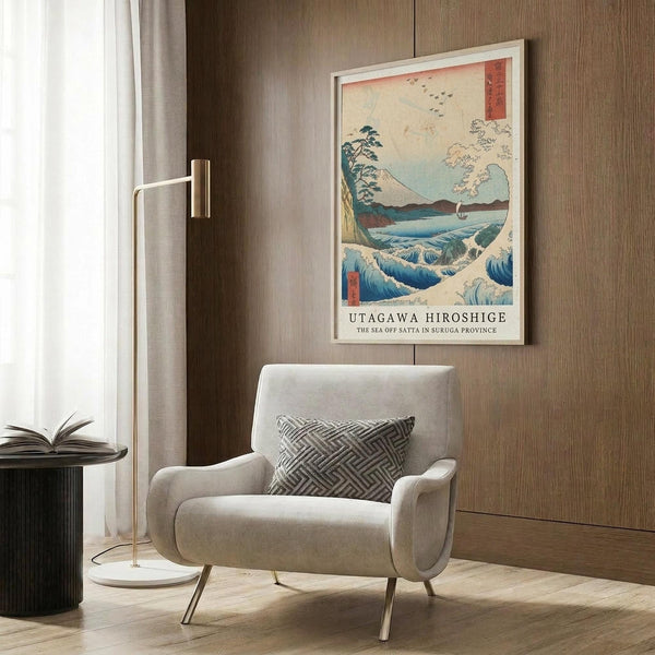 May include: Framed Japanese woodblock print featuring blue ocean waves, Mount Fuji, and birds in flight, displayed above a beige upholstered armchair with geometric pillow. Modern living room setting with brass floor lamp, dark round side table with open book, and wood-paneled wall. Print shows traditional ukiyo-e art style with vibrant blues, coral accents, and cream tones in white frame.