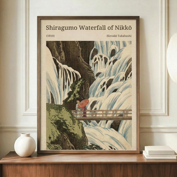 May include: Framed Japanese woodblock print reproduction showing a dramatic waterfall cascading over dark rocks with two figures holding a red umbrella standing on a wooden bridge. The artwork features traditional ukiyo-e style with flowing white and blue water against moss-covered cliffs. Displayed on a wooden sideboard next to a white ceramic vase and stacked books in a room with white paneled walls.