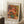 May include: Framed art print of impressionist-style roses in a blue vase, featuring soft pink and coral blooms with warm yellow and green background brushstrokes. The poster has "VASE OF ROSES" at top and "PIERRE AUGUSTE RENOIR 1890-1900" at bottom. Shows wooden frame leaning against white paneled wall on textured white rug, with coffee cup on wooden block and beige fabric visible.