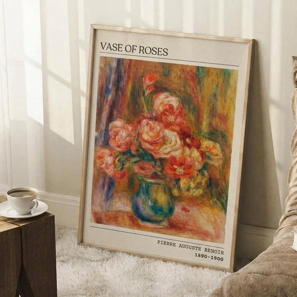 May include: Framed art print of impressionist-style roses in a blue vase, featuring soft pink and coral blooms with warm yellow and green background brushstrokes. The poster has "VASE OF ROSES" at top and "PIERRE AUGUSTE RENOIR 1890-1900" at bottom. Shows wooden frame leaning against white paneled wall on textured white rug, with coffee cup on wooden block and beige fabric visible.