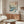 May include: Japanese woodblock print poster showing ocean waves in blue and white with Mount Fuji, trees, and birds in background, displayed in thin frame above wooden sideboard with sliding doors. Modern living room setting features white upholstered chair, glass oval coffee table, brass floor lamp, and neutral beige walls.