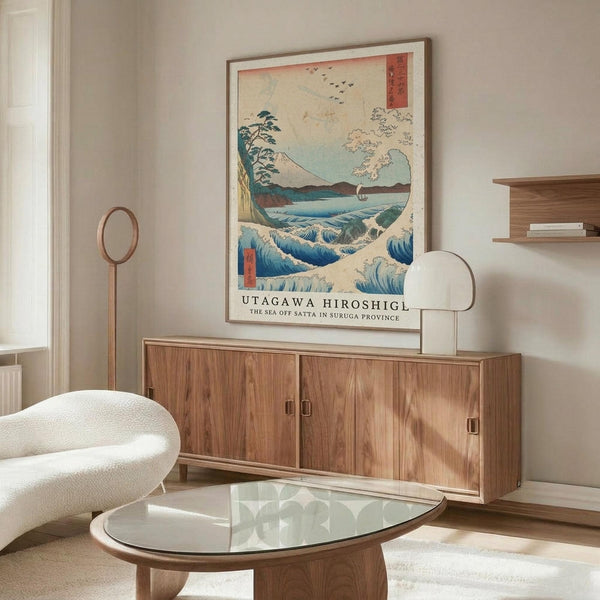 May include: Japanese woodblock print poster showing ocean waves in blue and white with Mount Fuji, trees, and birds in background, displayed in thin frame above wooden sideboard with sliding doors. Modern living room setting features white upholstered chair, glass oval coffee table, brass floor lamp, and neutral beige walls.