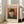 May include: A framed Van Gogh museum exhibition poster featuring a self-portrait of the artist wearing a dark fur hat and green coat while smoking a pipe, set against an orange-red background. The vintage-style poster has cream borders with museum text and is displayed in a light wood frame, leaning against a beige upholstered chair in a bright, minimalist room with white walls and large windows.