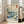 May include: Framed Japanese woodblock print reproduction displayed on hardwood floor beside beige sectional sofa. The artwork features dramatic blue ocean waves with white foam, Mount Fuji in background, traditional sailing vessel, and flying birds under cloudy sky. Text identifies artist Utagawa Hiroshige and location "The Sea off Satta in Suruga Province" below the scenic image. Light wooden frame complements bright, airy room with white walls and large windows.