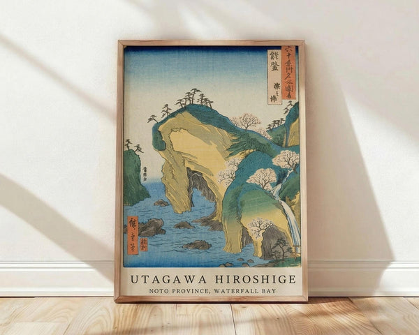 May include: Framed Japanese woodblock print showing dramatic coastal landscape with towering rock formations, waterfalls, and blue water. Features traditional ukiyo-e art style with green and yellow cliffs, cherry blossoms, and Japanese text in red cartouche. Natural wood frame displayed on light wooden floor against white wall with soft shadow patterns.