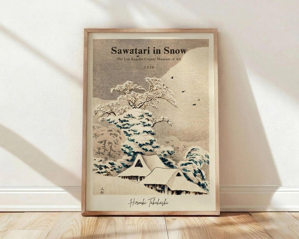 May include: Framed vintage Japanese woodblock print showing snow-covered traditional houses beneath bare winter trees with flying birds, displayed in light wood frame against white wall on wooden floor. Artwork features muted beige and blue tones with museum exhibition text at top and artist signature at bottom.