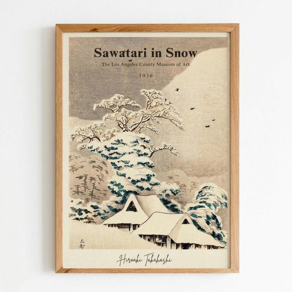 Japanese woodblock print style artwork featuring snow-covered houses with steep roofs nestled among bare winter trees. The scene shows rolling hills in muted beige and brown tones with dark blue-green foliage details. Small birds fly in the overcast sky. The vintage-style poster includes museum attribution text at the top and an artist signature at the bottom. Displayed in a light wood frame against a white wall.