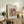 May include: Framed vintage-style art print featuring a tender mother and child painting in soft pastels, displayed on rustic wooden sideboard alongside fern plant, books, and ceramic vase in warm neutral living room setting with beige armchair and natural lighting.