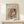 May include: Framed art print of Renoir's mother and child painting displayed on light wood console table against cream wall. Print shows woman in pink dress holding blonde toddler in red outfit, rendered in soft impressionist style with warm peachy tones. Small white wrapped gift with greenery sits beside the thin natural wood frame. Gentle shadows cast across minimal, neutral-toned room setting.