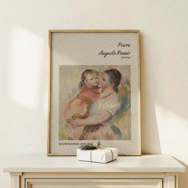 May include: Framed art print of Renoir's mother and child painting displayed on light wood console table against cream wall. Print shows woman in pink dress holding blonde toddler in red outfit, rendered in soft impressionist style with warm peachy tones. Small white wrapped gift with greenery sits beside the thin natural wood frame. Gentle shadows cast across minimal, neutral-toned room setting.