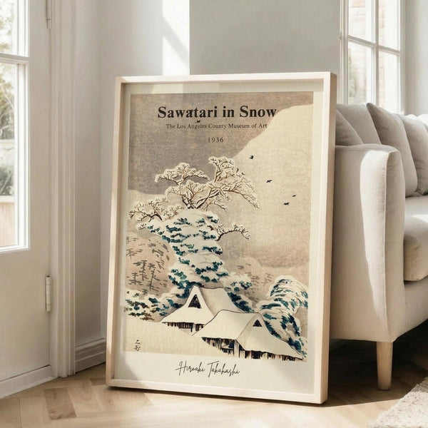 May include: Framed vintage Japanese woodblock print showing snow-covered houses beneath evergreen trees and bare branches, with flying birds against a beige textured background. The artwork features muted earth tones with blue-green accents and includes museum attribution text and artist signature. Displayed in light wooden frame leaning against beige armchair in bright room with white walls and windows.