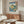 May include: Japanese woodblock print poster showing blue water, rocky cliffs, and flowering trees, displayed in thin wooden frame above walnut credenza. Modern living room setting with cream upholstered chair, glass-top oval coffee table, and brass floor lamp. Soft natural lighting creates shadows across white walls and polished floors.