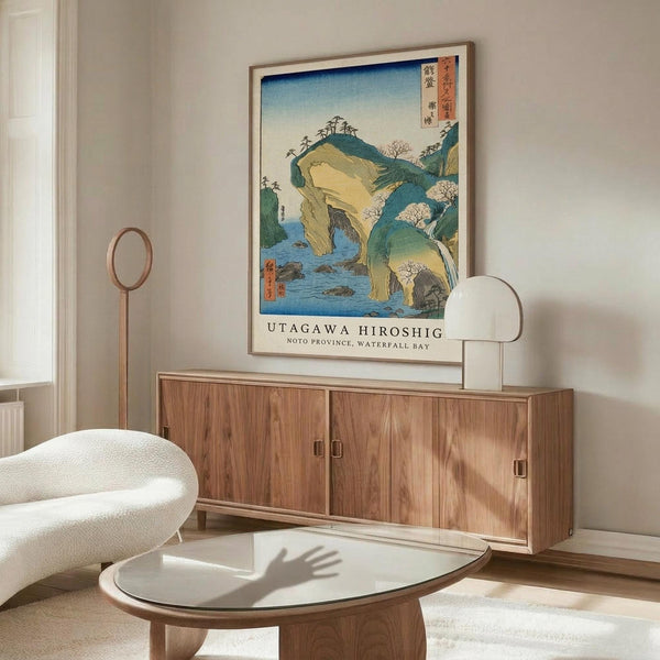 May include: Japanese woodblock print poster showing blue water, rocky cliffs, and flowering trees, displayed in thin wooden frame above walnut credenza. Modern living room setting with cream upholstered chair, glass-top oval coffee table, and brass floor lamp. Soft natural lighting creates shadows across white walls and polished floors.