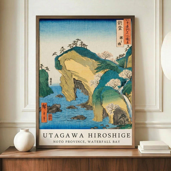 May include: Framed vintage Japanese woodblock print featuring dramatic coastal landscape with natural stone arch over blue water, green hills, and waterfall. Print displays traditional ukiyo-e art style with Japanese text cartouches. Frame sits on wooden credenza beside white ceramic vase and books in neutral-toned room with white walls and trim.