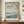 May include: Framed Japanese woodblock print showing cherry blossoms overhanging a serene river scene with Mount Fuji in the distance, small boats on water, and traditional landscape elements in soft blues, pinks, and greens. Print displayed on wooden credenza alongside white ceramic vase and stacked books in elegant room with white paneled walls and decorative lantern.