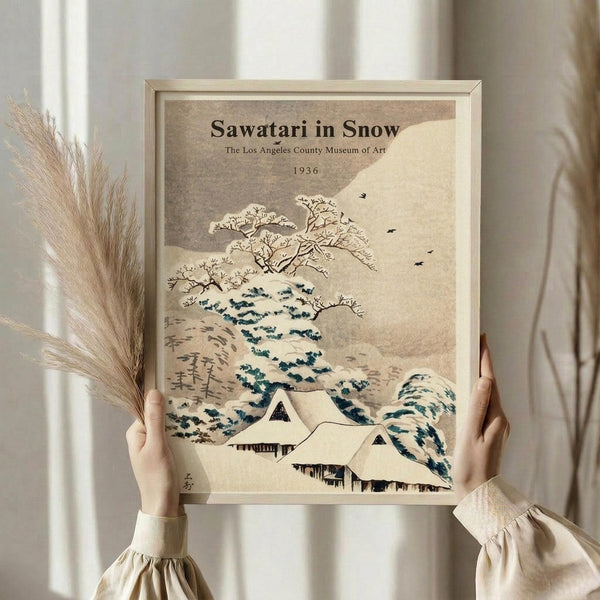 May include: Hands holding a framed vintage Japanese-style art print showing snow-covered houses beneath a large tree with bare branches, set against a beige winter landscape with small birds in flight. The artwork features muted earth tones and traditional Asian brushwork style. A bundle of dried wheat or pampas grass sits beside the frame against a neutral background with soft window lighting.