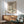 May include: Framed vintage-style Japanese winter landscape poster showing snow-covered houses beneath a large tree with blue water, displayed above a wood credenza with ceramic lamp in modern living room with mint green armchair and round coffee table, natural sunlight casting shadows on white wall