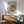 May include: Framed Renoir reproduction print showing a woman in soft pastels holding a small child, hanging on white wall above wooden sideboard with white ceramic lamp. Mid-century modern sage green armchair with wooden arms sits nearby in sunlit room with minimalist decor.