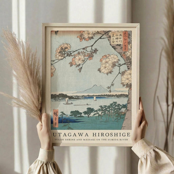 May include: Person in cream outfit holding framed Japanese woodblock print featuring cherry blossoms, Mount Fuji, and river scene with boats. Dried pampas grass visible to left. Print shows traditional ukiyo-e art style with pink blossoms overhead, blue water, green landscape, and distant mountain. Light wooden frame against neutral curtained background.