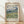 May include: Framed Van Gogh museum poster leaning against white wall on light wood floor with soft natural lighting and fabric shadows. The print features a swirling landscape with dramatic blue and white clouds above rolling green hills and golden wheat fields in Van Gogh's distinctive brushstroke style. Light wooden frame with cream matting displays the 1889 artwork with museum-style text below describing the vivid colors and composition.