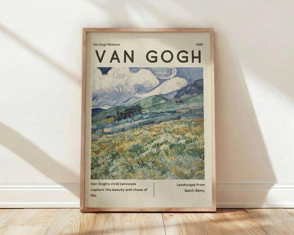 May include: Framed Van Gogh museum poster leaning against white wall on light wood floor with soft natural lighting and fabric shadows. The print features a swirling landscape with dramatic blue and white clouds above rolling green hills and golden wheat fields in Van Gogh's distinctive brushstroke style. Light wooden frame with cream matting displays the 1889 artwork with museum-style text below describing the vivid colors and composition.