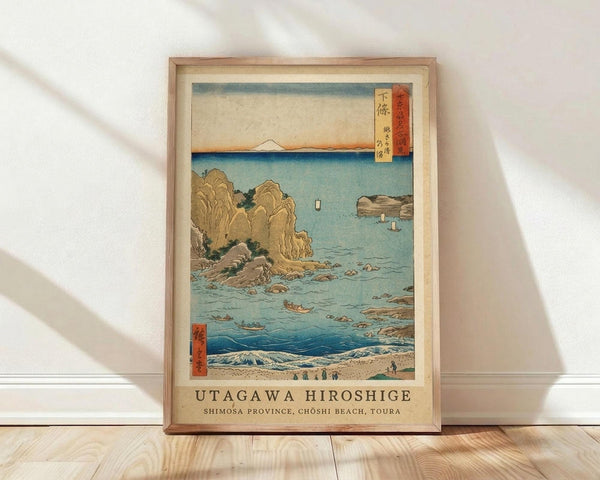 May include: Framed Japanese woodblock print reproduction showing a coastal scene with rocky cliffs, turquoise water, small boats, and Mount Fuji in the distance. The artwork features traditional ukiyo-e style with muted earth tones and blue hues, displayed in a light wood frame against white walls on wooden flooring with soft natural lighting casting shadows.