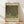 May include: Framed art print leaning against white wall on light wood floor with natural sunlight casting diagonal shadows. The poster features Gustav Klimt's impressionistic painting of dense green foliage and flowering rosebushes beneath large trees, with cream-colored text at top and bottom identifying the artwork and artist. The print has a thin natural wood frame and museum-style layout with beige borders.