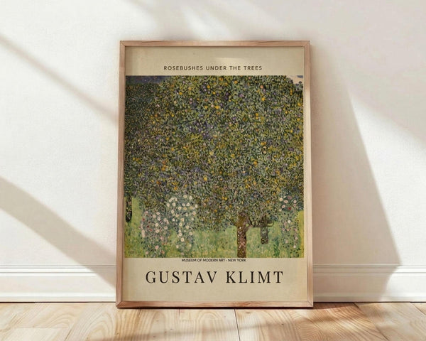 May include: Framed art print leaning against white wall on light wood floor with natural sunlight casting diagonal shadows. The poster features Gustav Klimt's impressionistic painting of dense green foliage and flowering rosebushes beneath large trees, with cream-colored text at top and bottom identifying the artwork and artist. The print has a thin natural wood frame and museum-style layout with beige borders.
