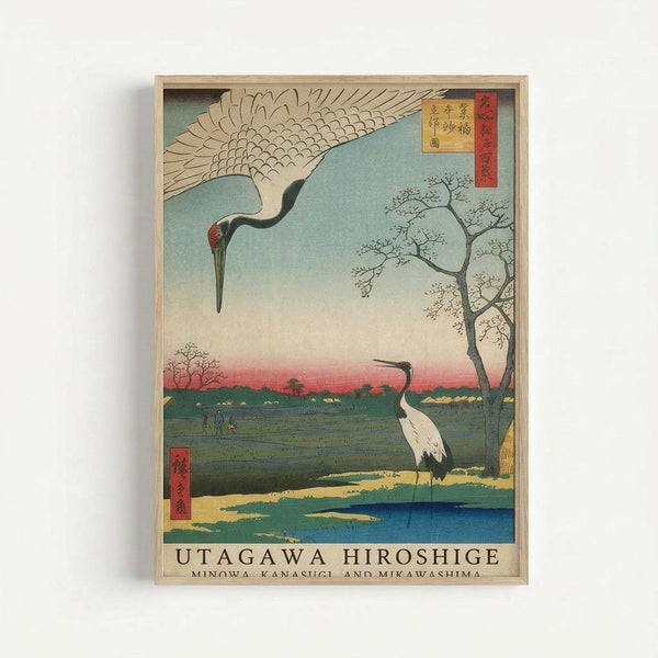 A framed Japanese woodblock print featuring two cranes in a serene landscape. One white crane with black wing tips flies overhead with outstretched wings, while another stands in shallow blue water below. The scene shows a pastoral setting with bare trees, green fields, and a gradient sky transitioning from blue to pink at the horizon. Red Japanese text cartouches appear in the upper right and lower left corners. The artwork displays the characteristic flat colors and clean lines of traditional
