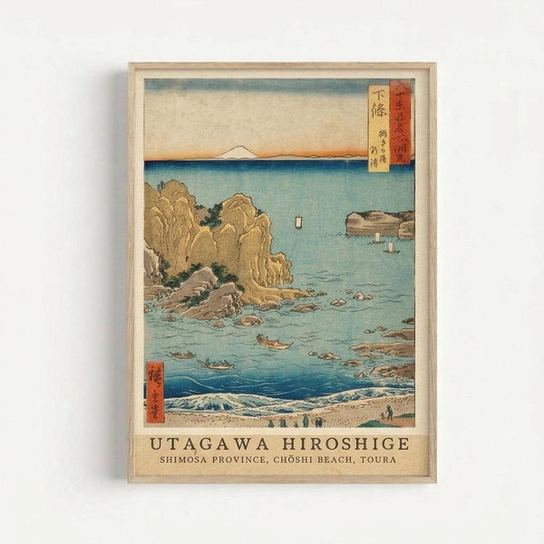 Framed Japanese woodblock print showing a coastal scene with rocky cliffs, turquoise waters, small boats, and waves in the foreground. Mount Fuji appears as a distant white peak on the horizon. The artwork features traditional ukiyo-e style with muted earth tones and blue-green water. Red Japanese text cartouches appear in the upper right and lower left corners. The print is displayed in a light wood frame against a white wall, with text below reading "UTAGAWA HIROSHIGE SHIMOSA PROVINCE, CHOSHI