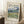 May include: Framed Van Gogh museum poster showing swirling landscape painting with dramatic blue sky, rolling green hills, and golden wheat field in foreground. Light wood frame leans against white paneled wall next to wooden side table with coffee cup and beige throw blanket on textured rug.