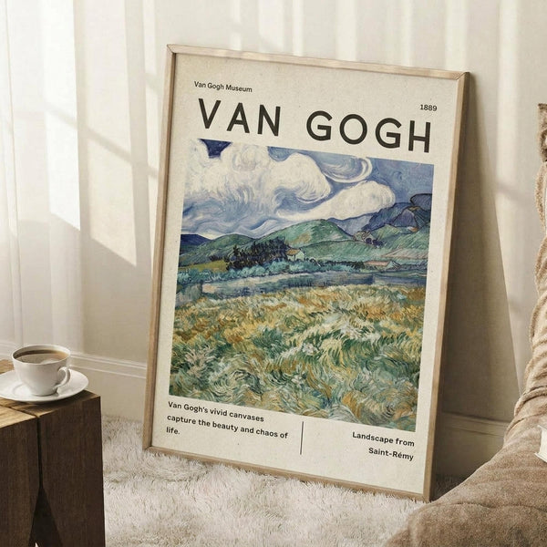 May include: Framed Van Gogh museum poster showing swirling landscape painting with dramatic blue sky, rolling green hills, and golden wheat field in foreground. Light wood frame leans against white paneled wall next to wooden side table with coffee cup and beige throw blanket on textured rug.