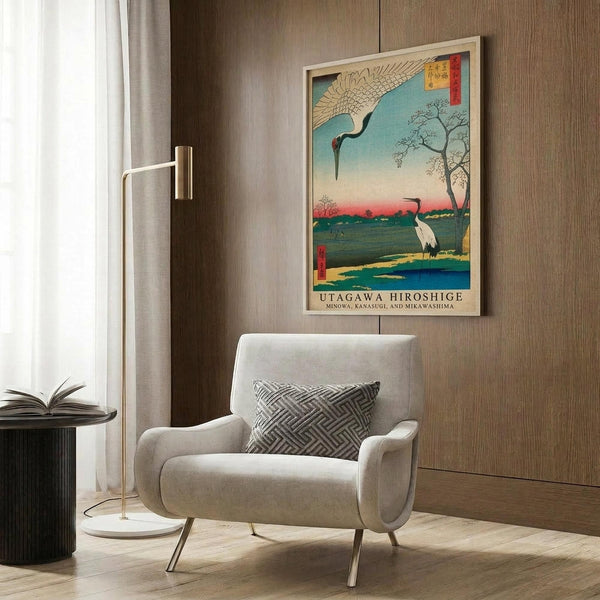 May include: Framed Japanese woodblock print featuring cranes in a marsh landscape with blue water, bare tree, and colorful sky, displayed on wood paneled wall above cream upholstered armchair with geometric pillow, brass floor lamp, and dark round side table with open book in modern living room setting.