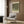 May include: Framed Gustav Klimt poster featuring a large tree with dense green foliage and white flowering rosebushes beneath, displayed on wood-paneled wall above beige upholstered armchair with geometric patterned pillow. Modern brass floor lamp and dark round side table with open book complete the neutral-toned reading nook with natural lighting from nearby window.