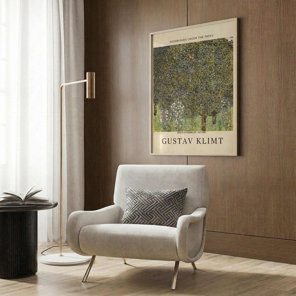 May include: Framed Gustav Klimt poster featuring a large tree with dense green foliage and white flowering rosebushes beneath, displayed on wood-paneled wall above beige upholstered armchair with geometric patterned pillow. Modern brass floor lamp and dark round side table with open book complete the neutral-toned reading nook with natural lighting from nearby window.