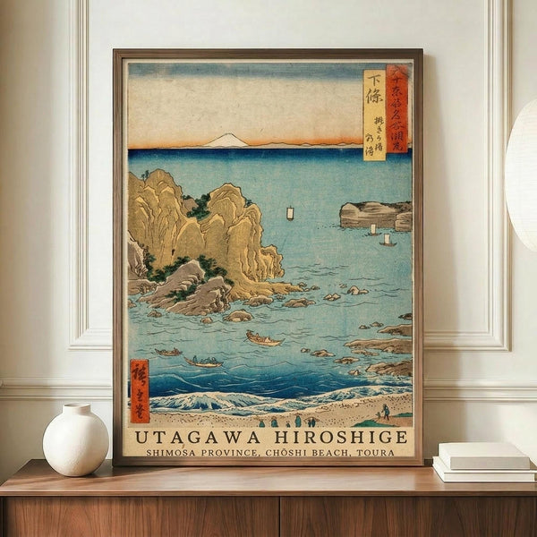 May include: Framed Japanese woodblock print depicting a coastal scene with rocky cliffs, turquoise waters, small boats, and Mount Fuji in the distance. The artwork features traditional ukiyo-e style with muted earth tones and blue gradients. Displayed on a wooden sideboard alongside a white ceramic vase and stack of books against white paneled walls in a modern interior setting.