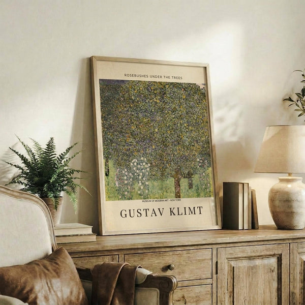 May include: Framed art print displaying Gustav Klimt's pointillist-style painting of dense green foliage and rosebushes with Museum of Modern Art New York attribution, positioned on rustic wooden sideboard alongside fern plant, ceramic vase, stacked books, and cream upholstered chair in neutral-toned living room setting