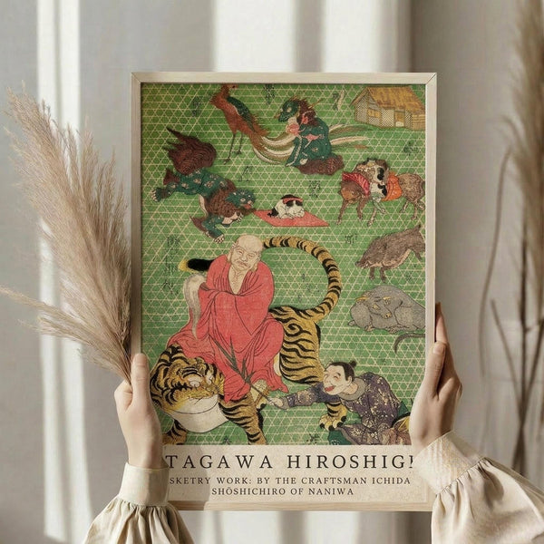 May include: Person in cream sweater holding framed Japanese art print featuring colorful illustration on green geometric background with monk in red robes sitting on striped tiger, surrounded by various animals and figures. Natural lighting with dried pampas grass visible to left side.